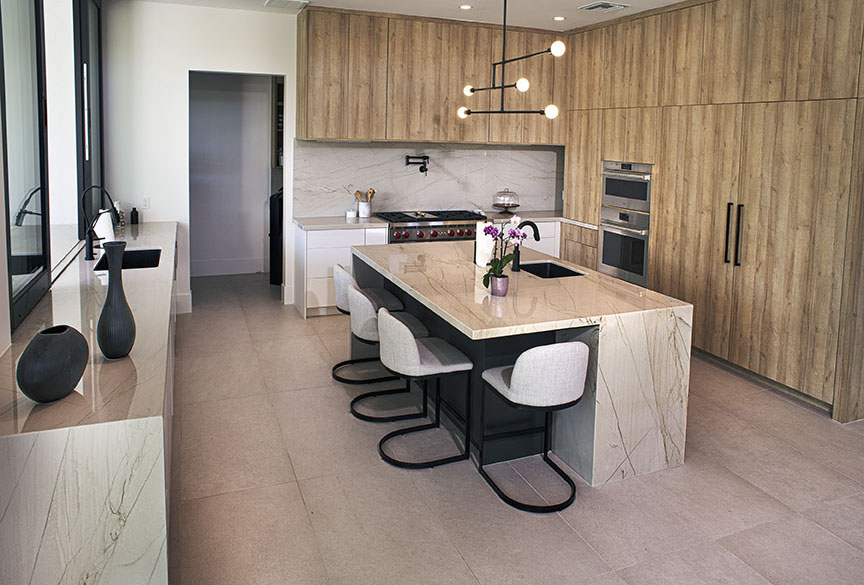 Interior with strobe lighting of a contemporary kitchen with large section light beige marble floors and lighter variegated marble counters and island with four seating positions and an oversized sink.